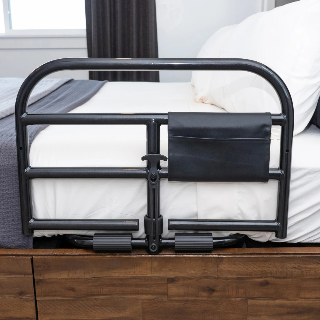 A Stander Prime Bariatric Safety Bed Rail in black metal with a small pocket is attached to the side of a bed with white bedding and pillows, providing added bedside safety next to a wooden bed frame.